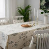 A dining table set with a cream tablecloth featuring a rustic fern print, surrounded by white wooden chairs and candles.