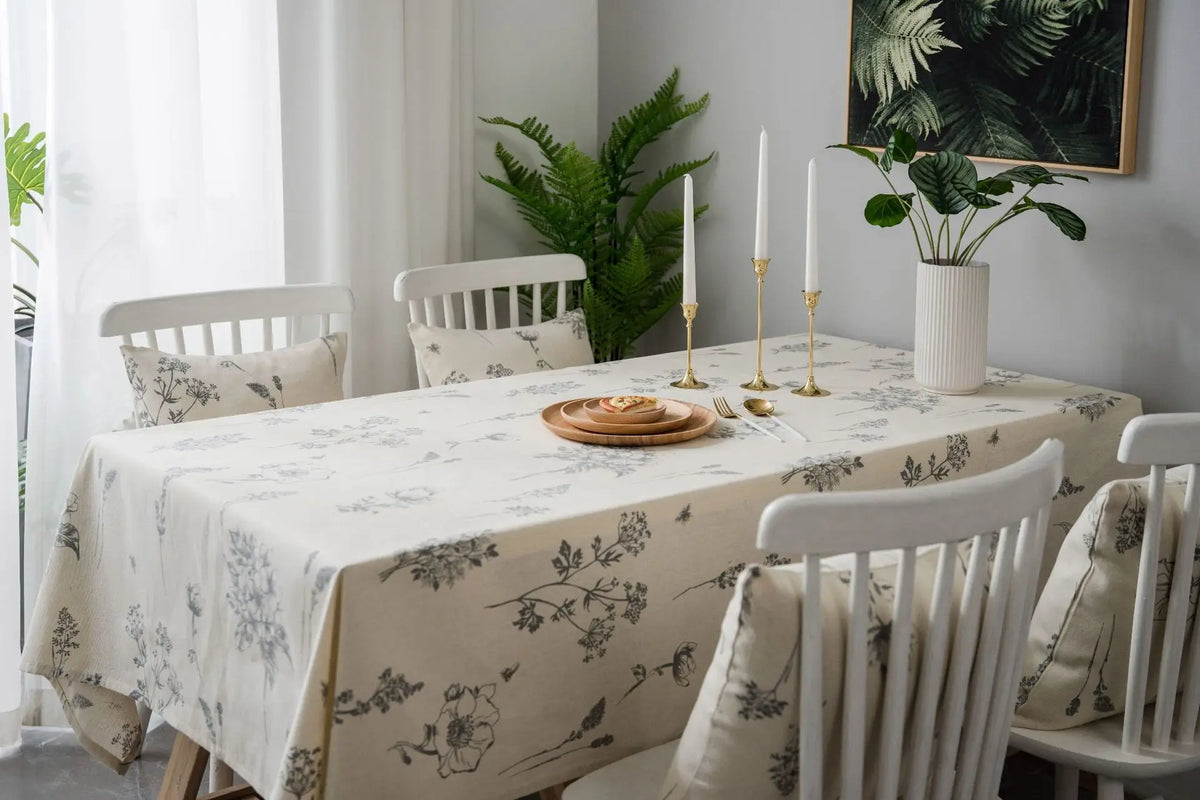 A dining table set with a cream tablecloth featuring a rustic fern print, surrounded by white wooden chairs and candles.