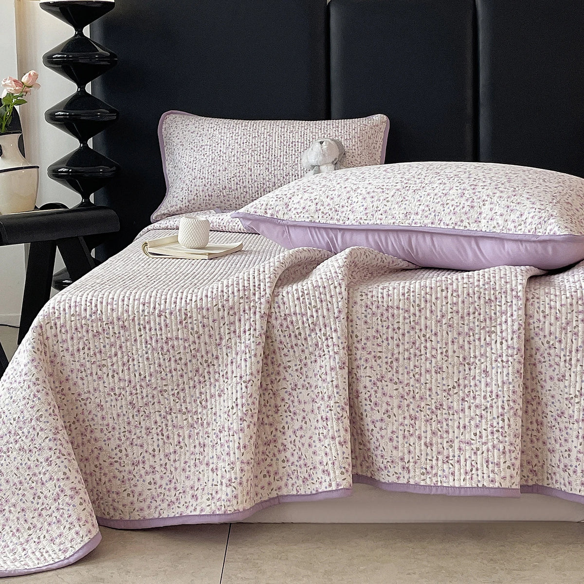 A floral quilted bedspread in soft pink and white patterns is draped over a dark headboard.
