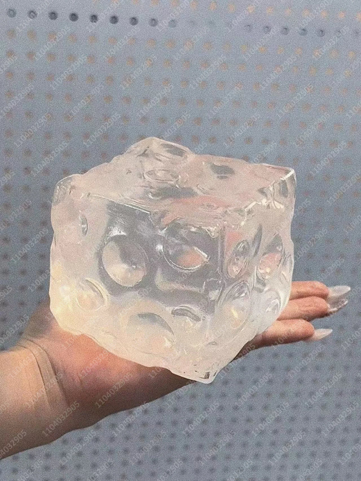 A hand holds a clear, frosted cube stress ball with various holes and shapes.