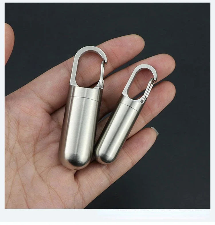 A hand holds a stainless steel pill capsule with a keychain loop.