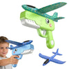 A green foam catapult toy shaped like a dinosaur with an attached airplane wing is shown next to a child's hand.