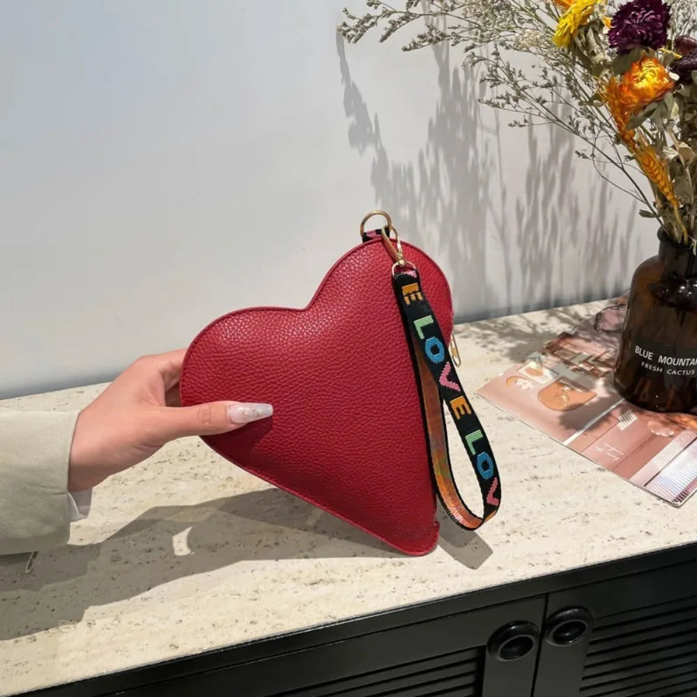 A red heart-shaped PU coin purse with a black and yellow strap is displayed on a light countertop.