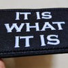 Black embroidered patch with white text reading IT IS WHAT IT IS