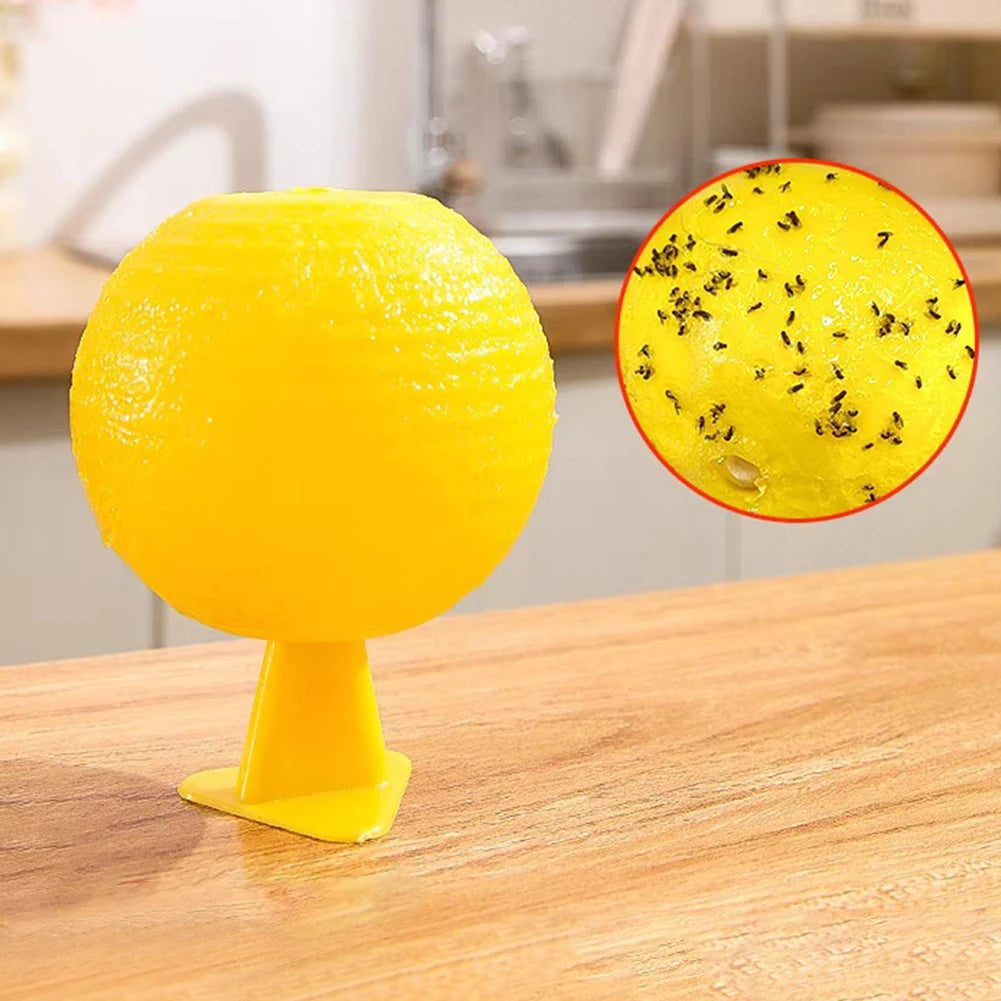 A bright yellow sticky fruit fly trap ball with black specks, displayed on a wooden surface.