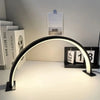 A half-moon LED arc desk lamp with a curved light illuminates a workspace next to framed art.