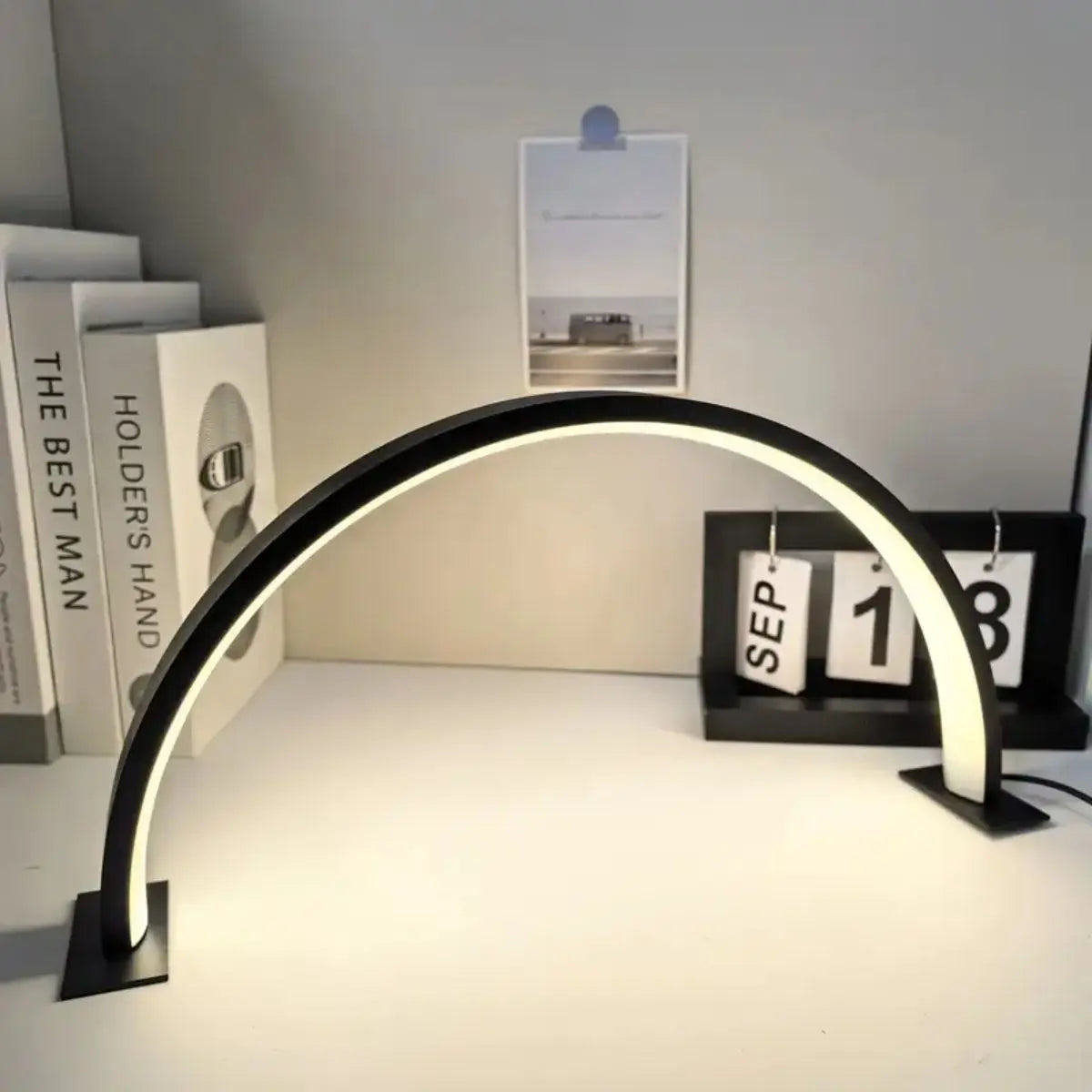 A half-moon LED arc desk lamp with a curved light illuminates a workspace next to framed art.