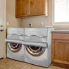 A pair of silver waterproof washing machine covers with roll edge designs are displayed next to a wooden cabinet.