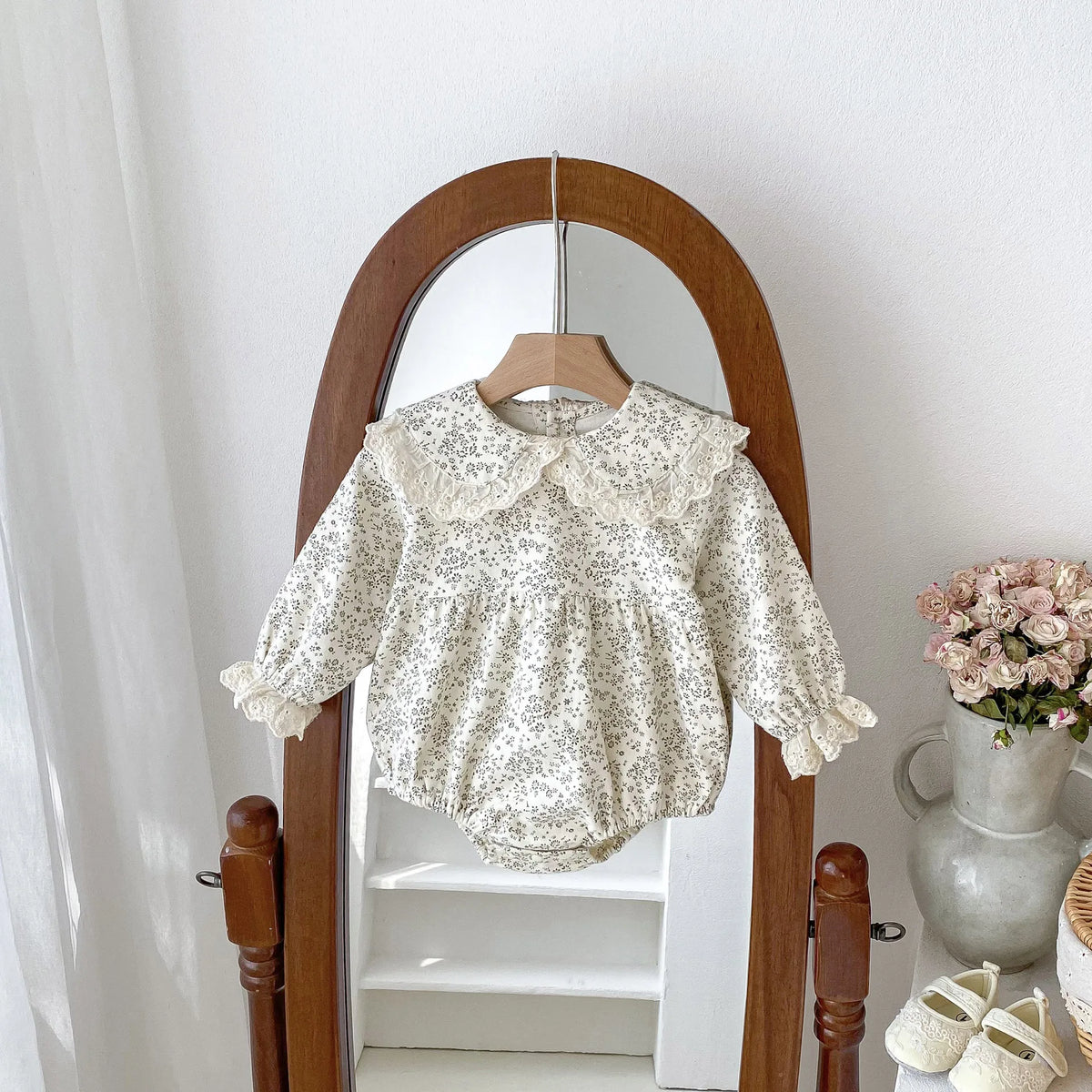 A white floral lace cotton romper for a baby girl hanging in a wooden-framed mirror.