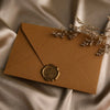 A kraft paper envelope sealed with a gold wax sticker and adorned with dried floral sprigs on a satin background.