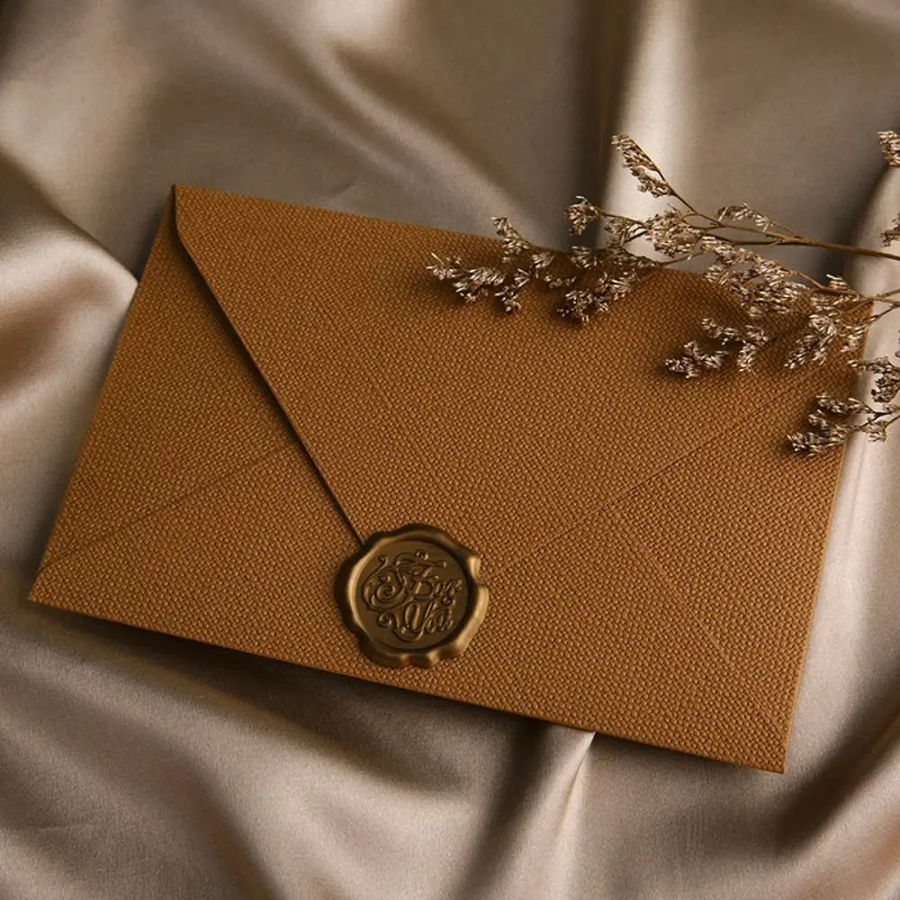 A kraft paper envelope sealed with a gold wax sticker and adorned with dried floral sprigs on a satin background.