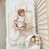 A baby wearing a beige outfit is lying on a portable diaper changing pad next to a wooden shelf.