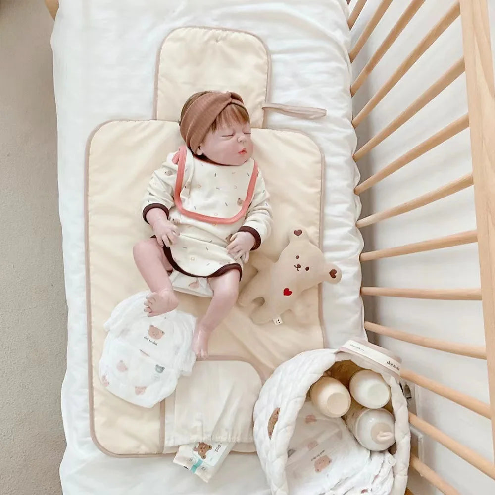 A baby wearing a beige outfit is lying on a portable diaper changing pad next to a wooden shelf.