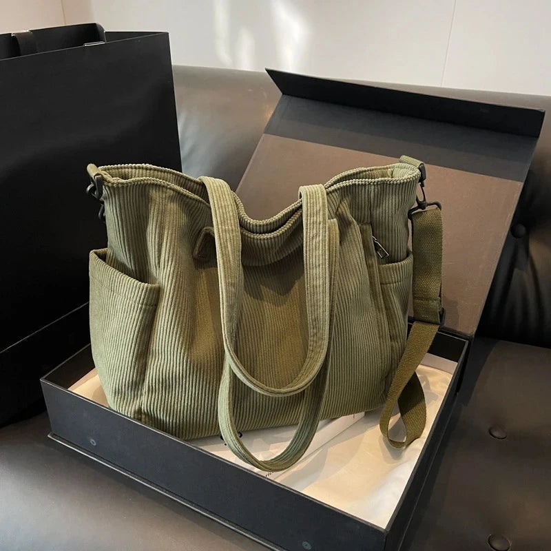 A olive green corduroy tote bag with an adjustable strap displayed in a black box.