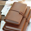 A stack of brown genuine leather refillable journals resting on a wooden surface with paper and tools.