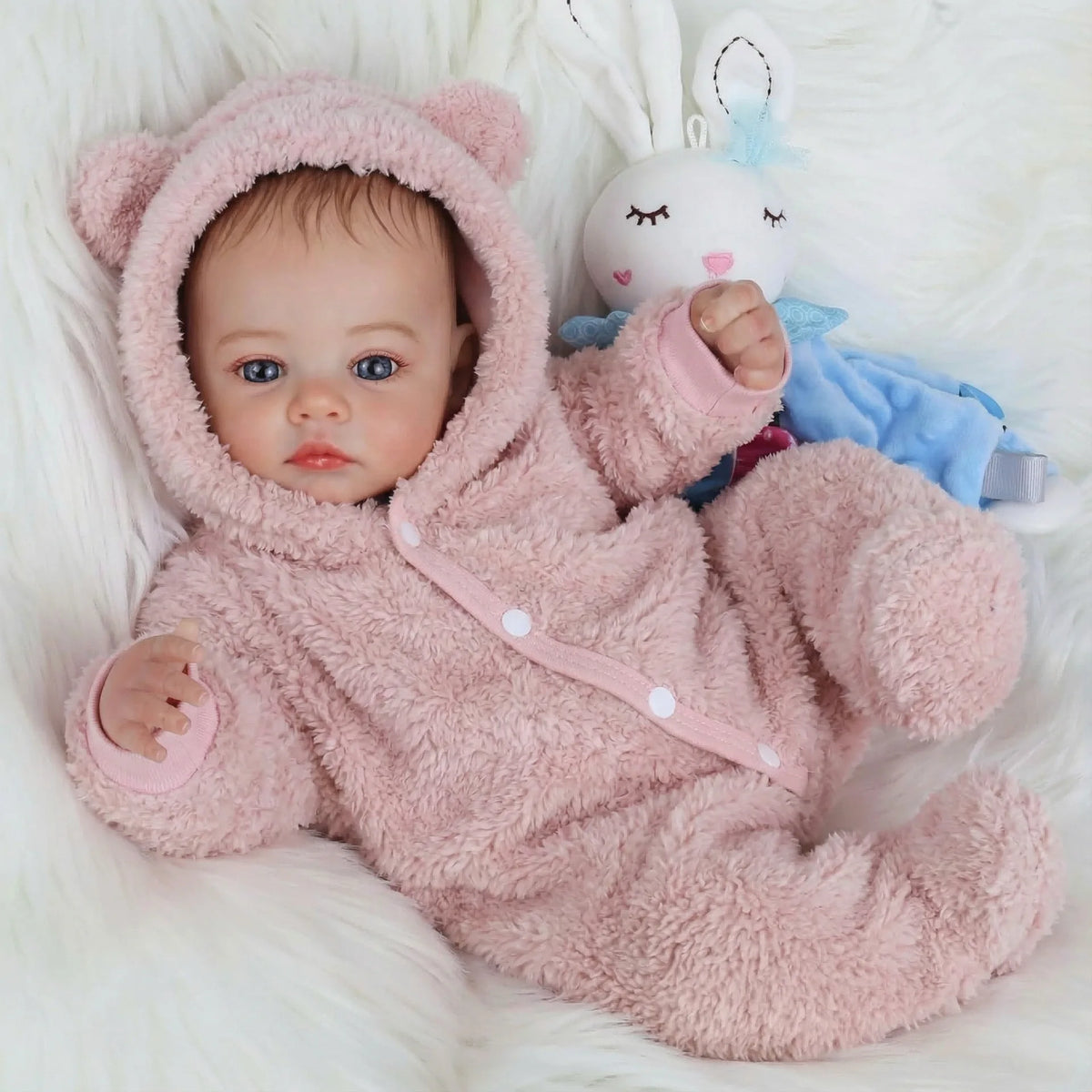 A newborn Reborn baby doll wearing a pink fuzzy hooded outfit lying on white fur.