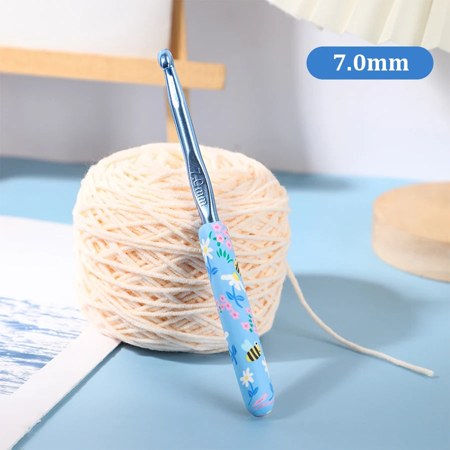A blue ergonomic crochet hook with a bee design is shown next to a ball of light-colored yarn.