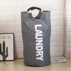 A large gray foldable laundry bag with a padded handle is displayed against a white tiled wall.