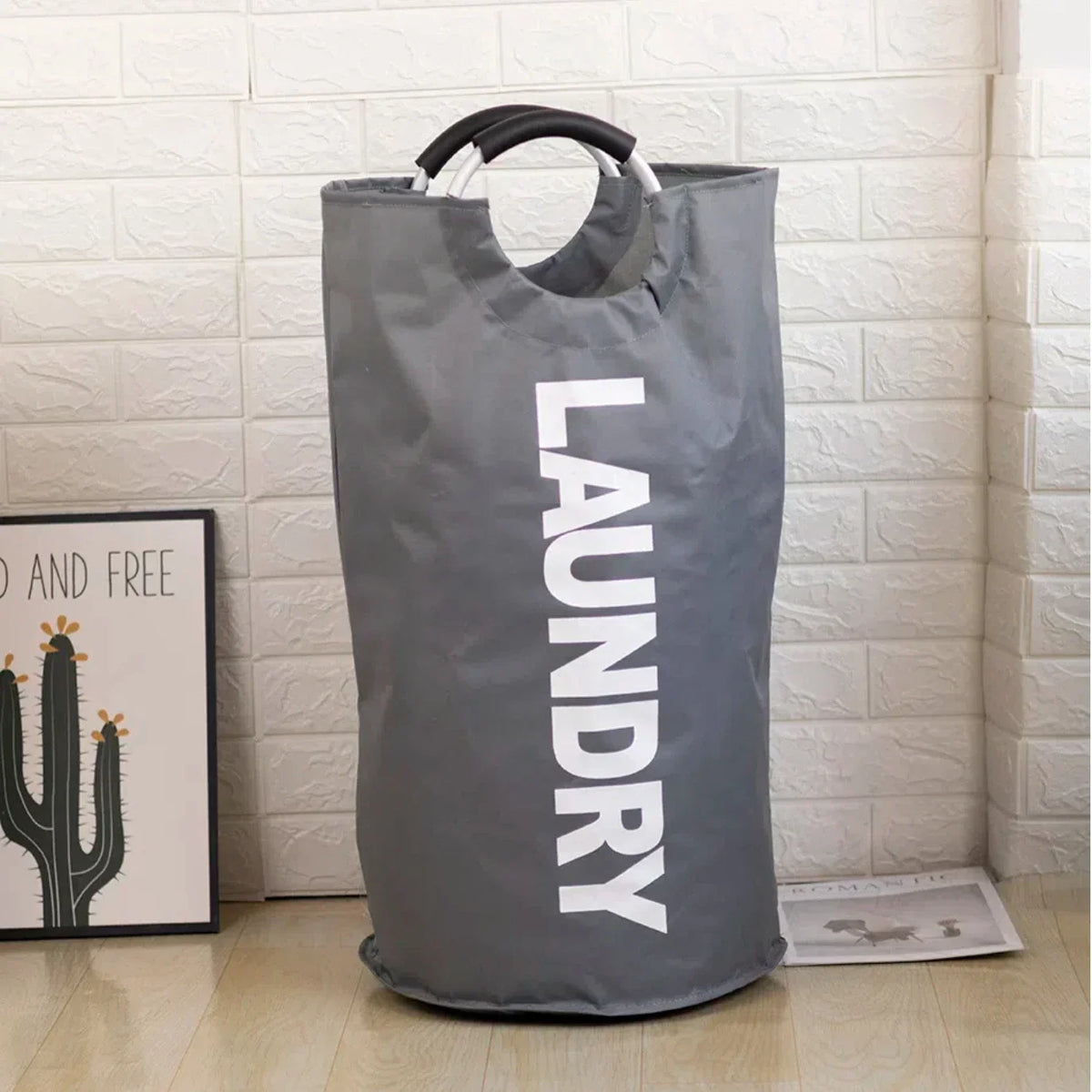 A large gray foldable laundry bag with a padded handle is displayed against a white tiled wall.