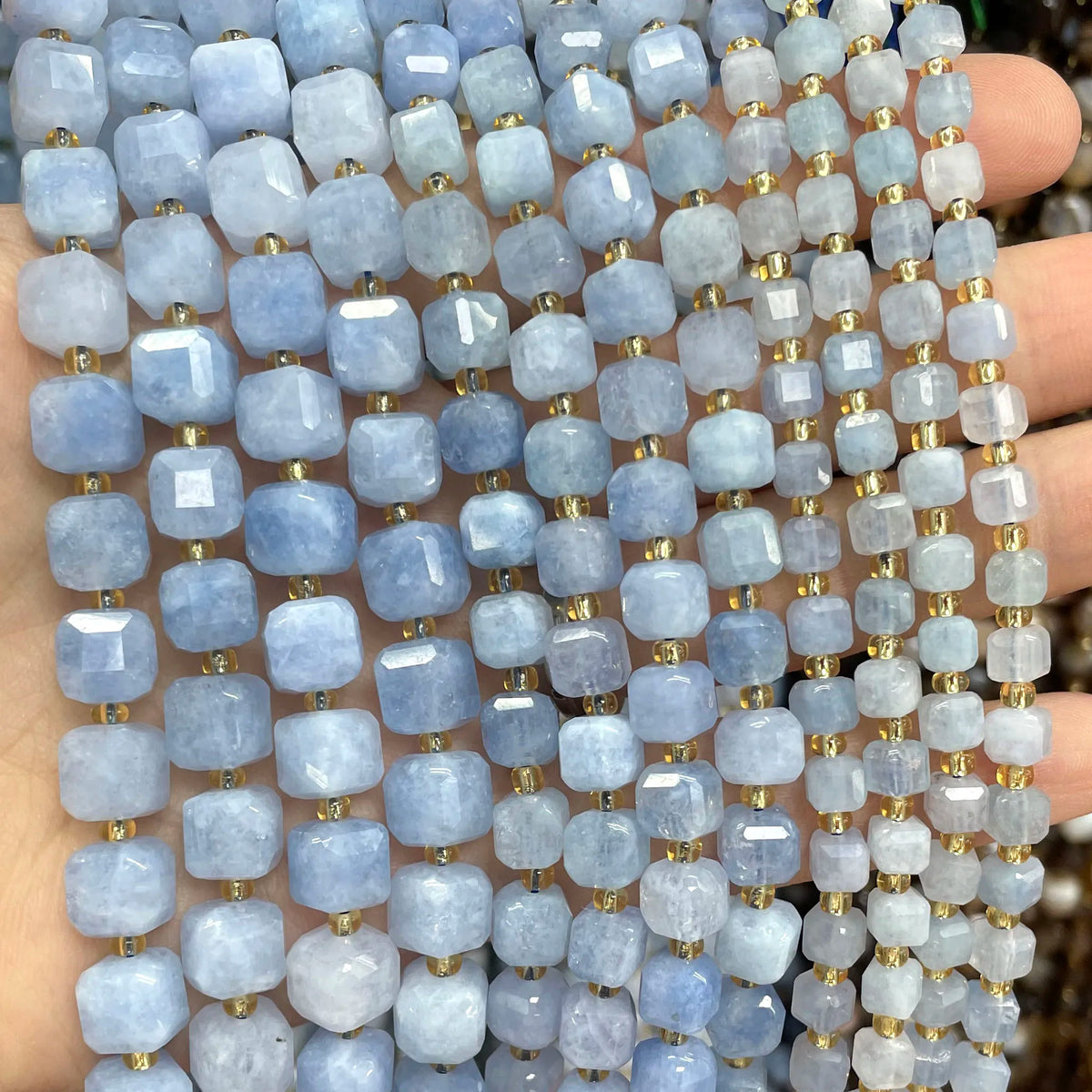A hand holds a strand of light blue and lavender square faceted natural stone beads for jewelry making.
