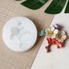Un moule en silicone blanc en forme de fleur est exposé sur un tapis tissé avec des feuilles vertes.
