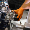 A hand wearing an orange nitrile glove is holding a metallic component of a machinery part.