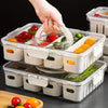 A hand is lifting the lid of a clear plastic portable snack container filled with colorful vegetables and fruits.