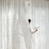 A person in a white dress is adjusting sheer, light-filtering lace curtains in a bright room.