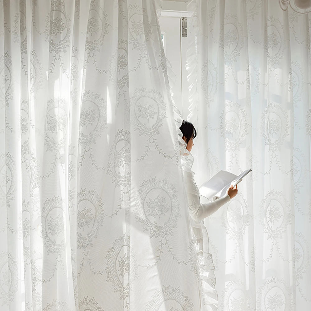 A person in a white dress is adjusting sheer, light-filtering lace curtains in a bright room.