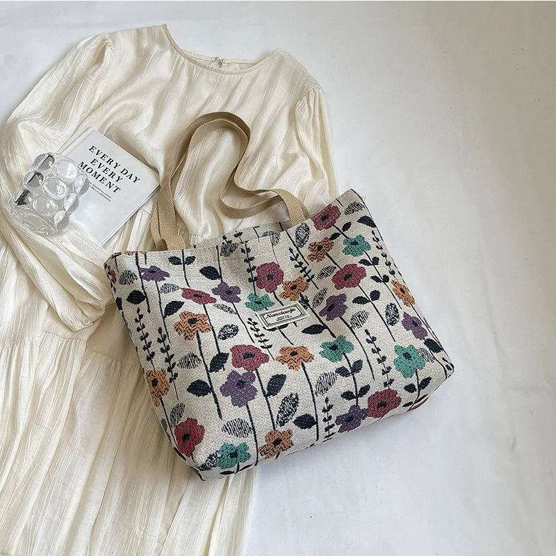 A beige corduroy tote bag with floral accents rests next to a white dress and a small glass object.
