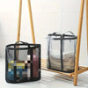 A foldable mesh laundry basket with a handle is displayed on a wooden rack against a white tiled background.