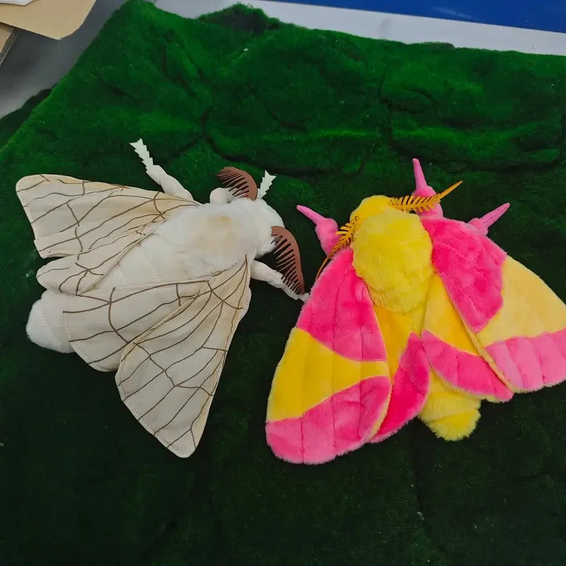 A plush toy of a Luna moth with white wings and a yellow and pink body set against a green fabric background.