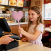 A young girl is holding a pink heart-shaped contactless payment wand in a cafe setting.