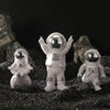 Three handcrafted astronaut figurines in white resin standing on dark gravel against a black background.