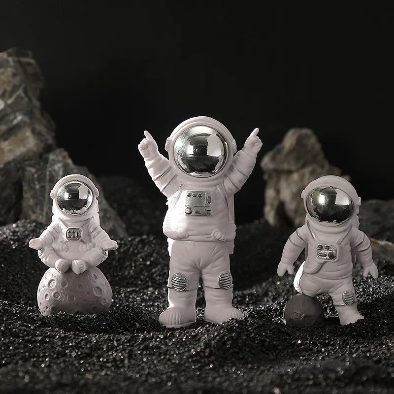 Three handcrafted astronaut figurines in white resin standing on dark gravel against a black background.