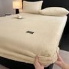 A beige plush mattress cover with an elastic edge is shown on a bed, next to a lamp and pillows.