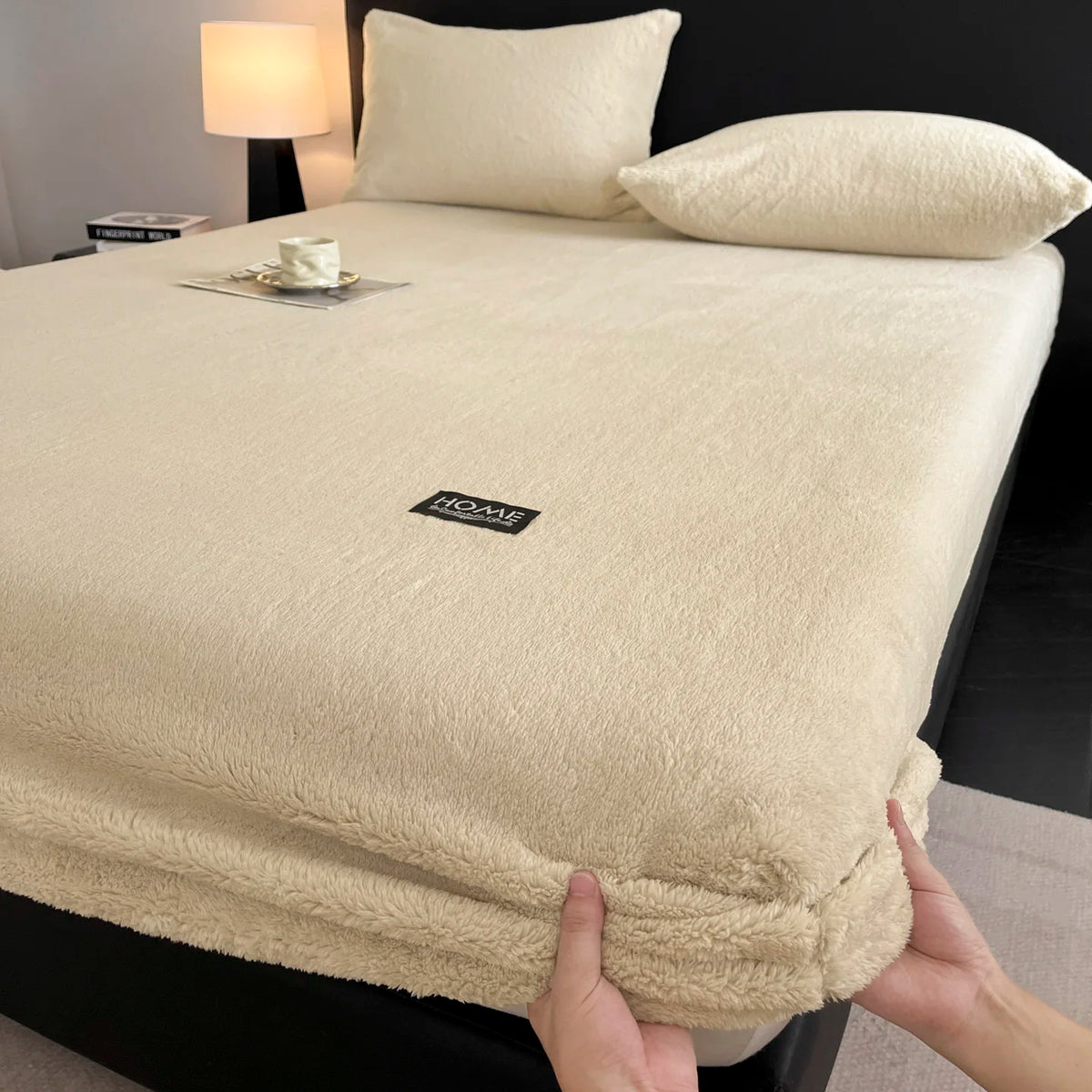 A beige plush mattress cover with an elastic edge is shown on a bed, next to a lamp and pillows.
