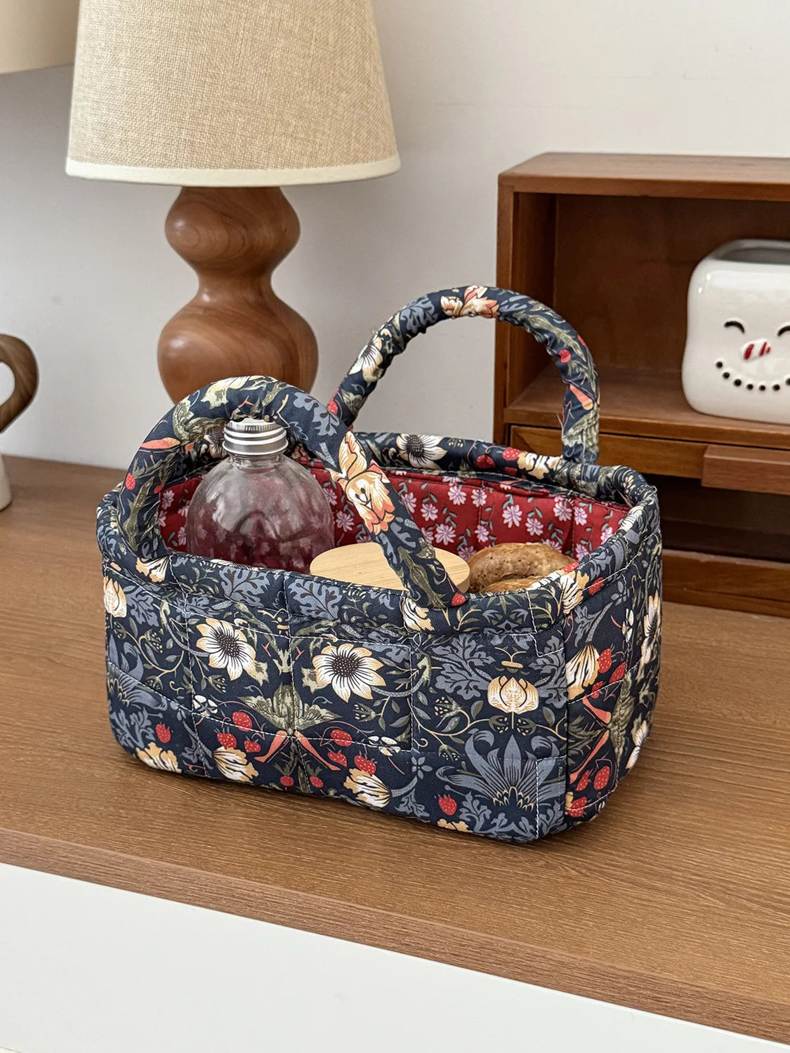 A dark floral patterned quilted fabric storage basket holding a glass bottle, resting on a wooden surface.