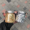 Two glass jars filled with metallic gold and silver foil flakes are held against a tiled background.