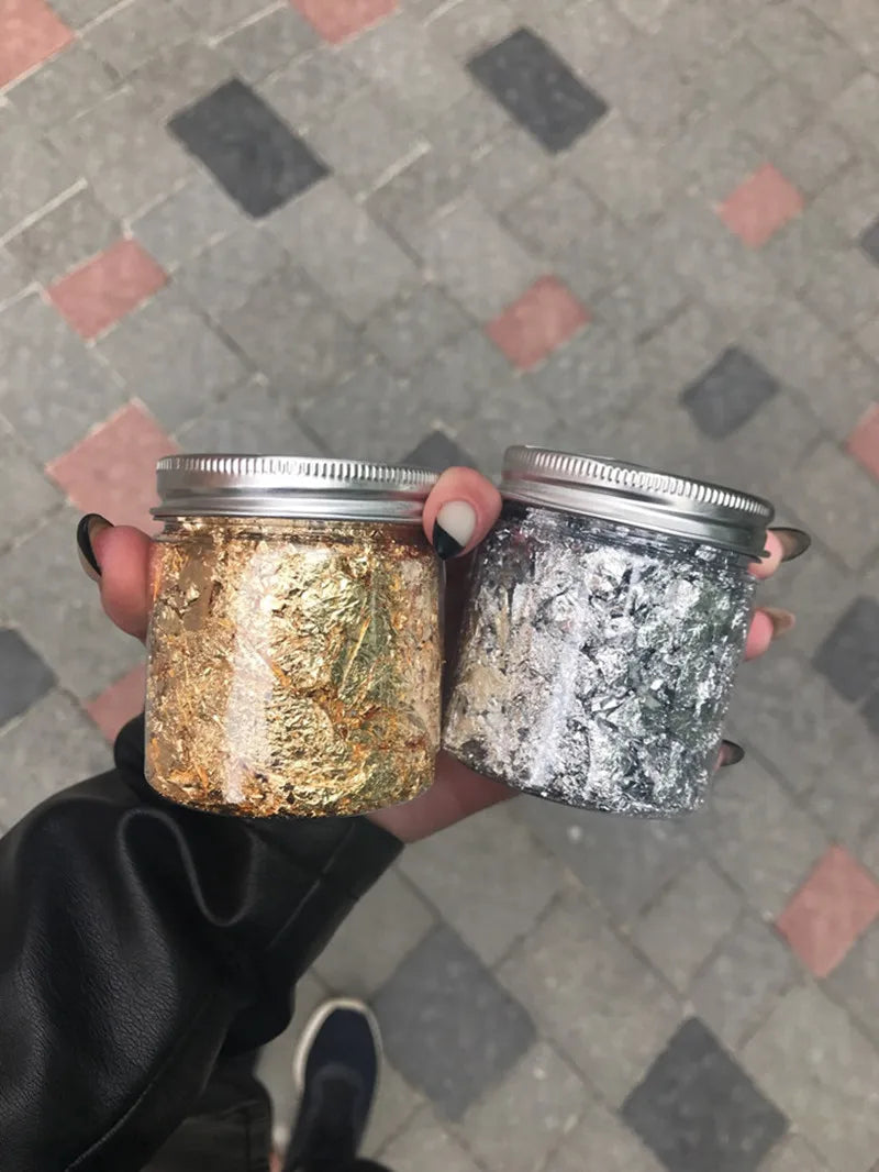 Two glass jars filled with metallic gold and silver foil flakes are held against a tiled background.