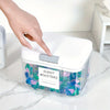 A hand places a silver pod holder into a clear plastic container filled with colorful laundry boosters.