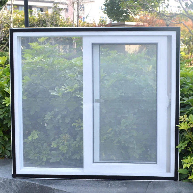 A white sliding window with a clear mesh screen installed in a black frame, set against green foliage.