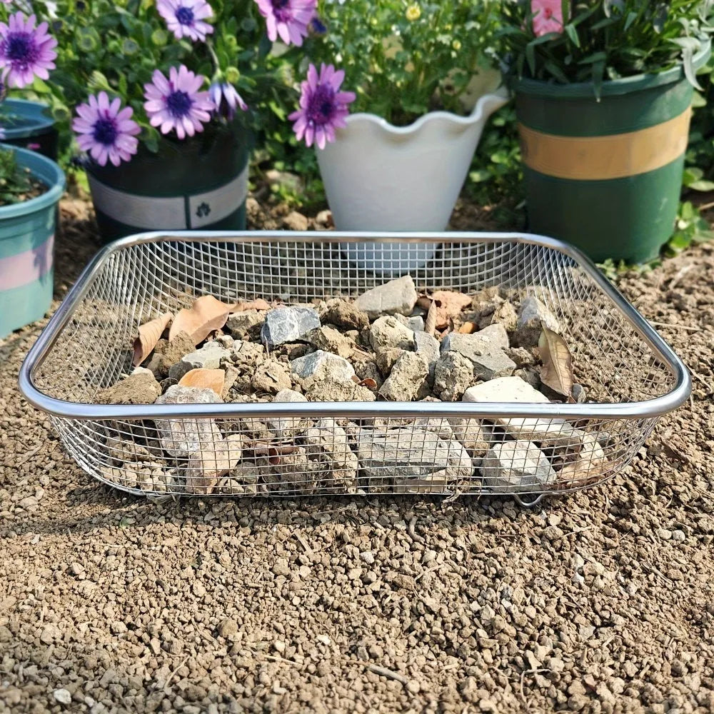 A stainless steel garden soil sieve filled with small rocks and soil, placed on dirt near potted plants.