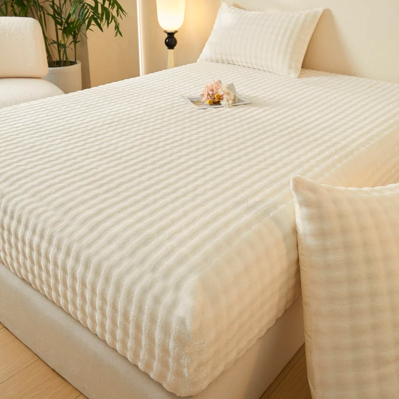 A white cozy faux fur fitted sheet is displayed on a bed with soft lighting and a potted plant in the background.