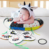 A baby wearing a cow hood is resting on a high contrast tummy time pillow with various shapes and patterns.