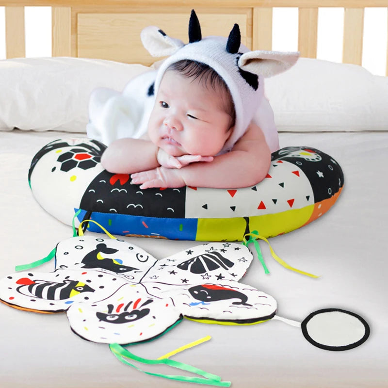 A baby wearing a cow hood is resting on a high contrast tummy time pillow with various shapes and patterns.