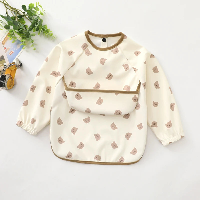 A cream waterproof long-sleeve smock for toddlers with a brown trim and small pink bear print.