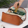 A hand is opening a spacious brown leather travel makeup organizer bag revealing various cosmetic bottles inside.