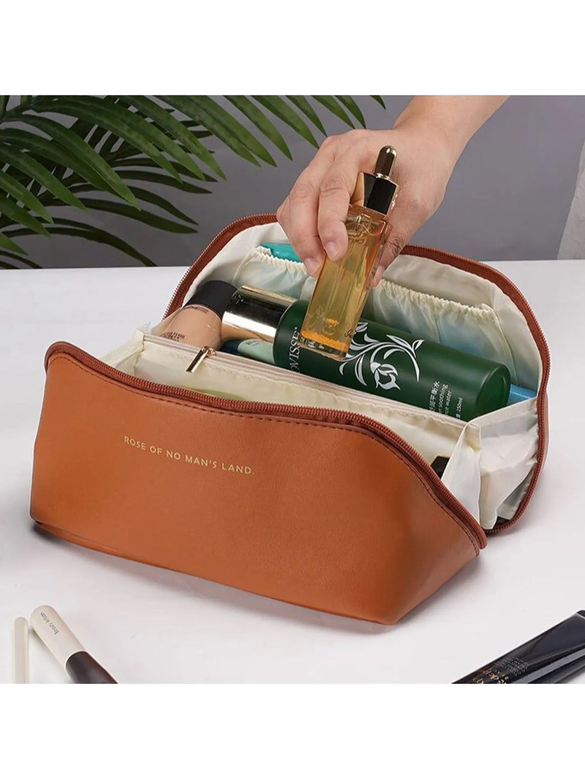 A hand is opening a spacious brown leather travel makeup organizer bag revealing various cosmetic bottles inside.