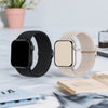A black and beige braided nylon watch band is shown next to a square Apple Watch, resting on a light surface with some papers